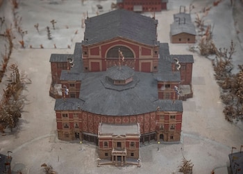 Modelo a escala de un edificio grande con techo oscuro y paredes rojas, rodeado de árboles miniatura y edificaciones menores, sobre una base clara