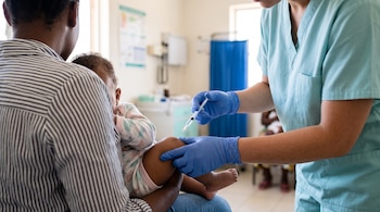 Una enfermera con guantes azules aplica una inyección en la pierna de un bebé sostenido por su madre dentro de un consultorio médico.