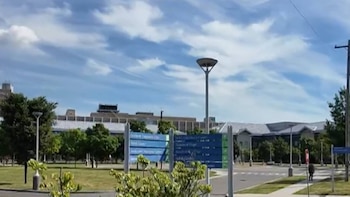 Cielo azul con nubes blancas, edificios modernos, árboles frondosos, farolas de diseño, calle pavimentada, aceras y un cartel indicador de direcciones
