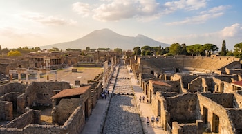 Vista aérea de las ruinas de Pompeya con edificios de piedra y calles adoquinadas bajo un cielo parcialmente nublado, y el Monte Vesubio en la distancia.