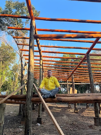 Un hombre viste sudadera amarilla y pantalón gris, sentado con las piernas cruzadas sobre un tronco horizontal que forma parte de una estructura de madera