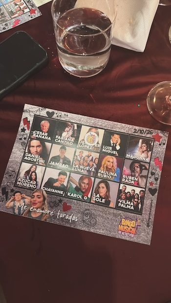 Cartón de juego con fotos de cantantes, la inscripción Bingo Musical, un vaso con agua y un teléfono sobre una mesa