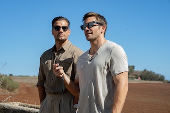 Dos hombres, Guy Ritchie y Jake Gyllenhaal, con gafas de sol, de pie al aire libre bajo un cielo azul claro, con un campo arado de fondo