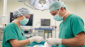 Dos cirujanos, hombre y mujer, con uniformes verdes, gorros, mascarillas y guantes, manipulan instrumental médico en un quirófano con monitores.