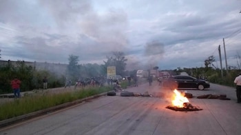 Fuego y neumáticos quemados sobre el asfalto de una carretera con personas, automóviles y camiones detenidos al fondo