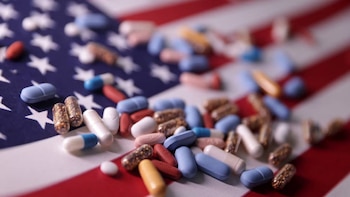 Primer plano de múltiples píldoras y cápsulas de diversos colores y tamaños, como azules, rojas, blancas y transparentes, distribuidas sobre una bandera de Estados Unidos desenfocada