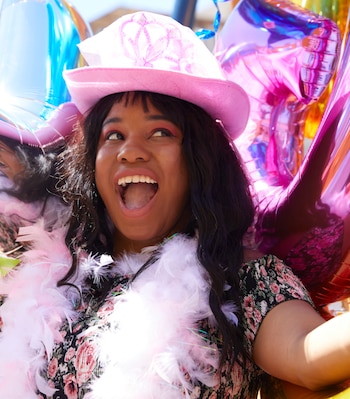 Una mujer con sombrero rosa, una boa blanca y vestimenta floral sonríe junto a varios globos metálicos de colores