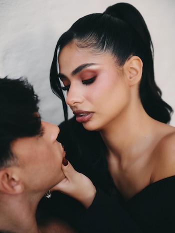 Primer plano de una mujer con maquillaje dramático tocando el mentón de un hombre, ambos con expresiones serias, fondo claro y desenfocado