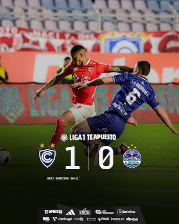 Dos futbolistas en acción durante un partido, uno con camiseta roja y otro con camiseta azul, disputando el balón. En la parte inferior, un marcador digital muestra '1-0'