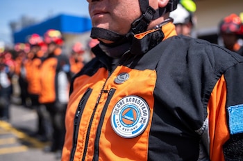 Hombre con uniforme naranja y negro, correa de casco y parche con los logos de GNE Acreditado, CONRED, USAR Mediano y Guatemala. Otros uniformados al fondo