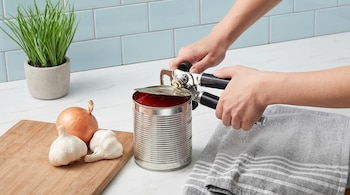 Manos abriendo una lata de salsa de tomate con un abrelatas. Sobre una mesada blanca hay cebolla, ajo, una tabla de madera y una toalla gris.