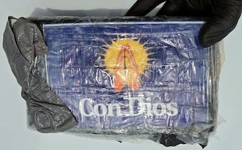 Un paquete rectangular envuelto en plástico transparente con cinta adhesiva gris y una etiqueta azul con unas manos rezando y el texto Con Dios