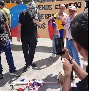 Grupo de personas en un espacio exterior, algunas sostienen una bandera de Venezuela, una pancarta amarilla con texto, dos cámaras, y trozos de tela de banderas en el suelo