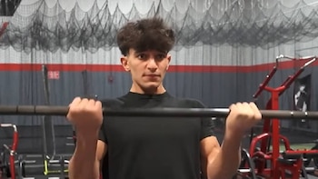 Hombre joven con cabello rizado y camiseta negra sostiene una barra a la altura del pecho en un gimnasio. Se observan redes en el techo y maquinaria roja al fondo