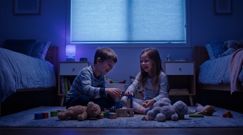 Dos niños sentados en una alfombra jugando con bloques de madera. Hay dos camas, dos mesas de noche y una lámpara encendida con luz azul.