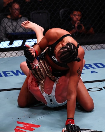 Dos luchadoras forcejean sobre la lona en un octágono, con manchas de sangre en el cabello y la espalda de la atleta situada en la parte inferior