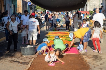 Un hombre con gorra azul crea una alfombra de aserrín colorida en el suelo, con diseños de un sol radiante y dos estructuras arquitectónicas coloniales, en una calle empedrada