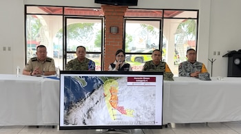 Cinco funcionarios sentados ante una mesa frente a una pantalla que exhibe el mapa de trayectoria del huracán Simon de categoría cuatro.