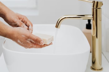 Unas manos con espuma sujetan una pastilla de jabón bajo el chorro de agua de un grifo dorado en un lavabo blanco