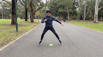 Un hombre con ropa deportiva salta de forma lateral sobre un cono verde en un camino asfaltado rodeado de árboles.