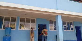Dos hombres frente a un edificio azul. Uno lleva un pulverizador de mochila. El otro usa chaleco y sombrero. Hay ventanas de marco blanco y barriles azules