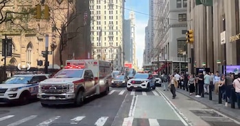 Una calle urbana con una ambulancia blanca y roja Ford y un SUV de policía Ford con luces de emergencia. Peatones en la acera y edificios altos al fondo