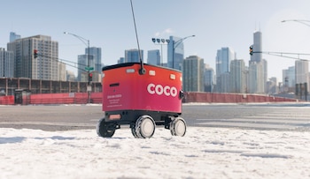 Un robot de delivery rojo y rosa con el logo 'COCO' y cuatro ruedas, parado en una calle con nieve en primer plano, con rascacielos urbanos al fondo