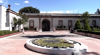 Reloj circular en el piso de un patio pavimentado ante un edificio blanco de estilo colonial con arcos y jardineras.