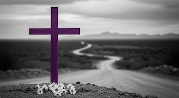 Cruz de madera morada con un ramillete de margaritas blancas sobre tierra. Al fondo, una carretera sinuosa en blanco y negro y montañas.