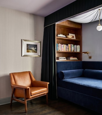 Un rincón de habitación con una estantería de madera llena de libros, un sofá azul oscuro, un sillón de cuero marrón y un cuadro en la pared.