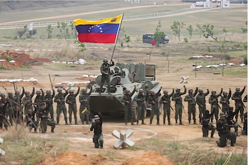Militares venezolanos, uniformados con cascos, levantan el puño en un campo. Un soldado sobre un vehículo blindado iza la bandera de Venezuela