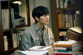 Choi Hyun-Wook, un joven con cabello oscuro y camisa gris, mira pensativo a la derecha mientras está sentado en un escritorio con libros y una estantería al fondo