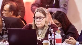 Mujer caucásica con cabello rubio y gafas, vestida con chaqueta burdeos y cuello alto, mirando una laptop negra sobre una mesa.
