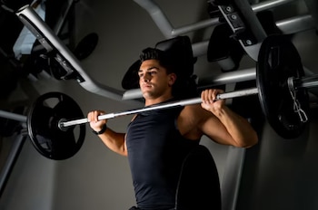 Un hombre joven vestido con camiseta de tirantes negra levanta una barra con discos de pesas. Se observa el equipo de gimnasio al fondo