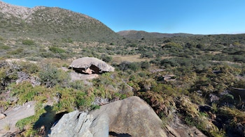 Un amplio paisaje montañoso con colinas cubiertas de vegetación rala y matorrales. Grandes rocas grises y marrones se ven en primer plano, con un cielo azul claro