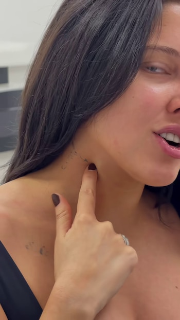 Una mujer de cabello oscuro señala con el dedo índice una marca tenue de tinta en el lado derecho de su cuello