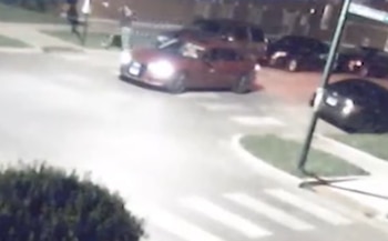 Toma nocturna de cámara de seguridad con un automóvil rojo de luces encendidas en una calle y varios vehículos estacionados