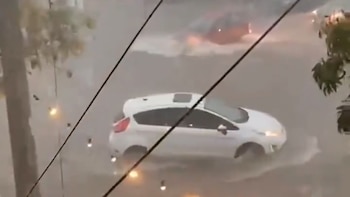 Dos automóviles, uno blanco y otro rojo, transitan por una calle completamente inundada bajo una fuerte lluvia, con cables eléctricos cruzando el encuadre
