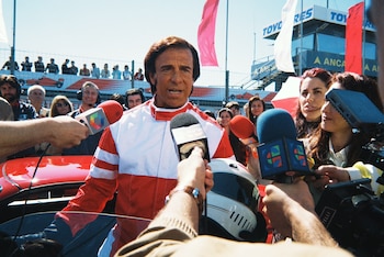 Un hombre con traje de piloto rojo y blanco sostiene un casco y habla ante varios micrófonos de prensa cerca de un automóvil deportivo