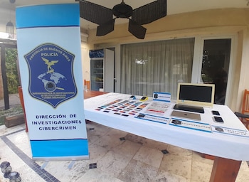 Mesa con pruebas incautadas por Cibercrimen, incluyendo tarjetas, documentos, computadoras y teléfonos, junto a un banner de la Policía de Buenos Aires