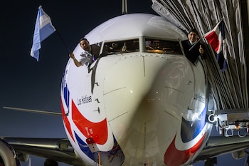 Dos hombres, probablemente pilotos, asoman por las ventanas de la cabina de un avión blanco por la noche, uno con la bandera argentina y otro con la dominicana