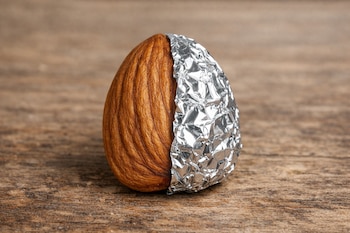 Una almendra entera se presenta sobre una superficie de madera, con una sección de su cuerpo cubierta por papel de aluminio arrugado y brillante.