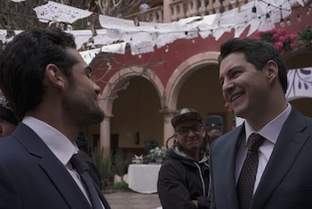 Dos hombres en trajes oscuros conversan y sonríen al aire libre, con el actor Carlos Gatica riendo a la derecha. Detrás, hay arcos rojos y papel picado