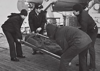 Fotografía en blanco y negro de cuatro hombres en abrigo oscuro que cargan a una persona cubierta en una camilla sobre un barco