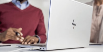 Persona con suéter y camisa interactuando con una laptop blanca HP, conectada con un cable, sobre una mesa. Fondo desenfocado con otra persona