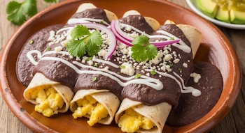 Primer plano de tres enfrijoladas con huevo revuelto, bañadas en salsa de frijol, con crema, queso, cebolla morada y cilantro, en un plato de barro. Fondo borroso con aguacate.