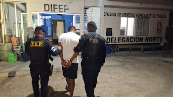 Dos agentes de la Policía Nacional Civil, vestidos de negro, escoltan a un hombre esposado con camiseta blanca y pantalón corto oscuro, frente a una estación policial