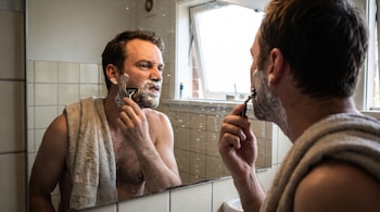 Hombre con toalla al hombro se afeita la barba con espuma y maquinilla, su reflejo visible en un espejo de baño salpicado de gotas cerca de una ventana.