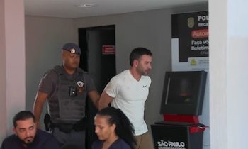 Un policía con uniforme gris escolta a un hombre con camiseta blanca y manos esposadas en la espalda dentro de un edificio oficial