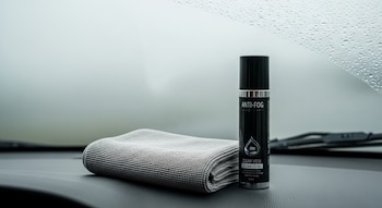 Un paño de microfibra gris y un envase negro de producto antiempañante sobre un tablero oscuro de vehículo, con un parabrisas con gotas de agua.